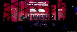 Pod podświetloną na czerwono ścianą z napisem Jubileuszowa gala EuroPower na krzesłach siedzi sześciu mężczyzn w garniturach i kobieta w czarnej marynarce i zielonej sukience, przed nimi zaciemniona sala.