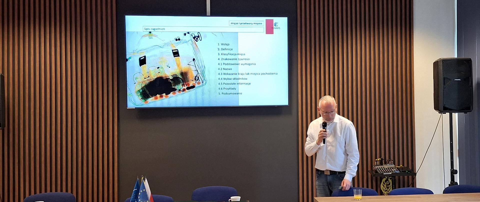 Jarosław Tymiński w trakcie wykładu. W prawym ręku trzyma mikrofon. Nad nim duży ekran monitora wyświetlającego slajd z treścią wykładu oraz zdjęciem poglądowym - zdjęciem prześwietlonej RTG torby.