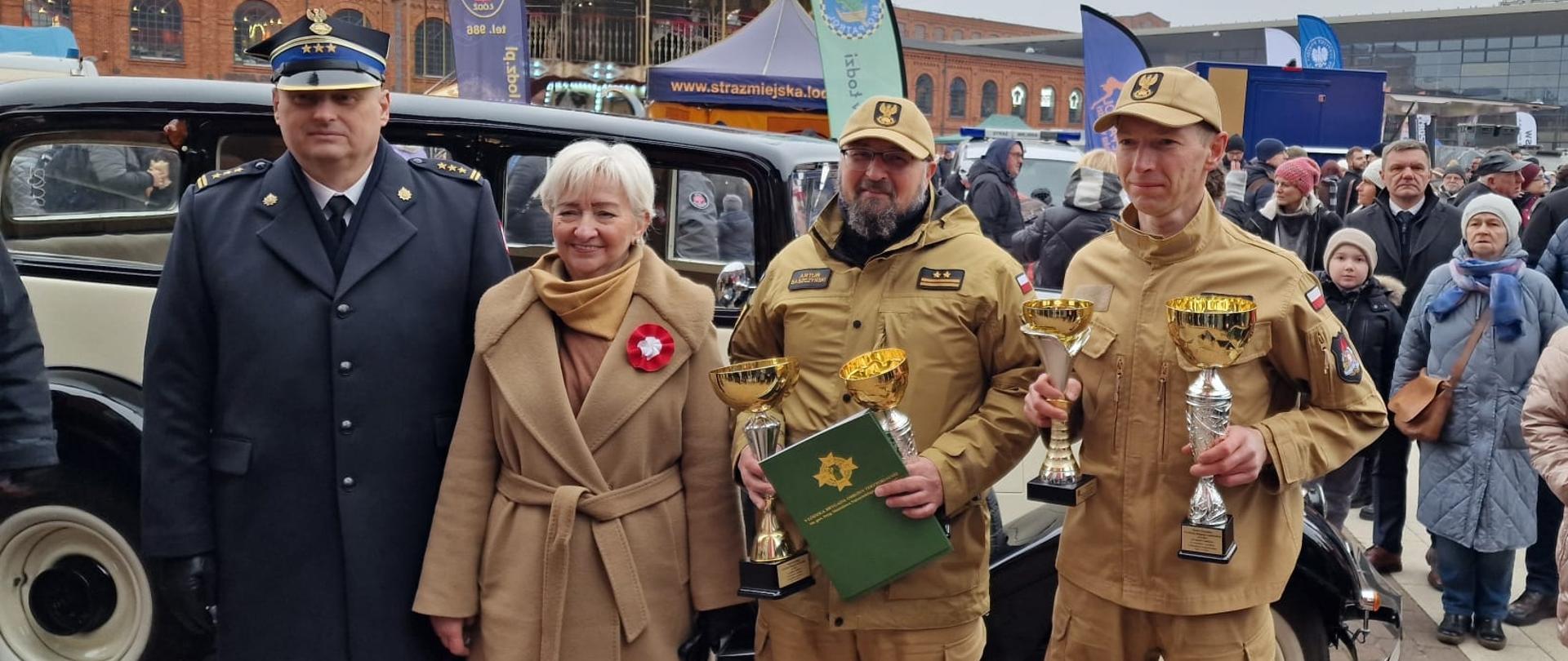 od lewej starszy brygadier Piotr Rudecki w płaszczu służbowym , Dorota Ryl Wojewódzka Łódzki w beżowym płaszczu, dwóch strażaków w mundurach służbowych w rękach trzymają puchary, wszyscy pozują do zdjęcia w tle zabytkowy samochód i ludzie