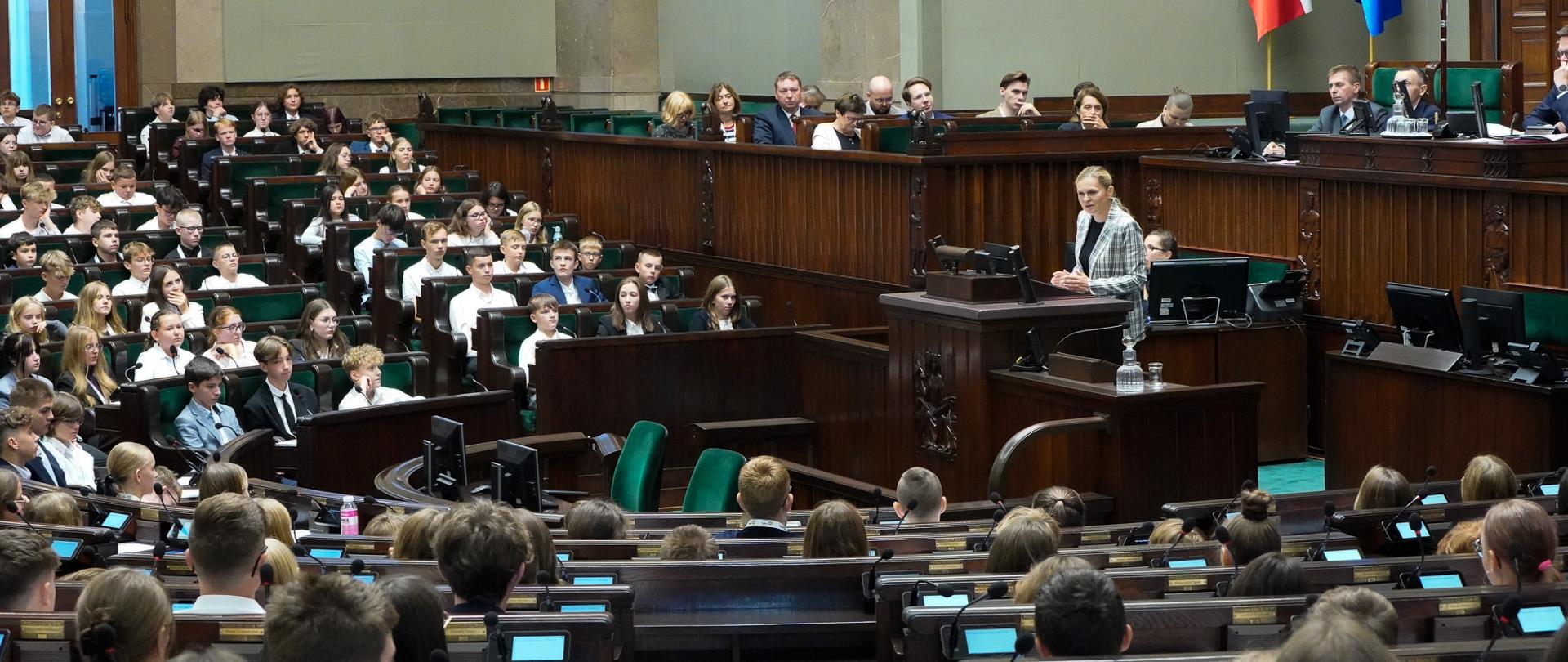 Widok z boku na sejmową salę obrad. W ławach poselskich siedzą dzieci i młodzież. Na mównicy przemawia minister Barbara Nowacka.