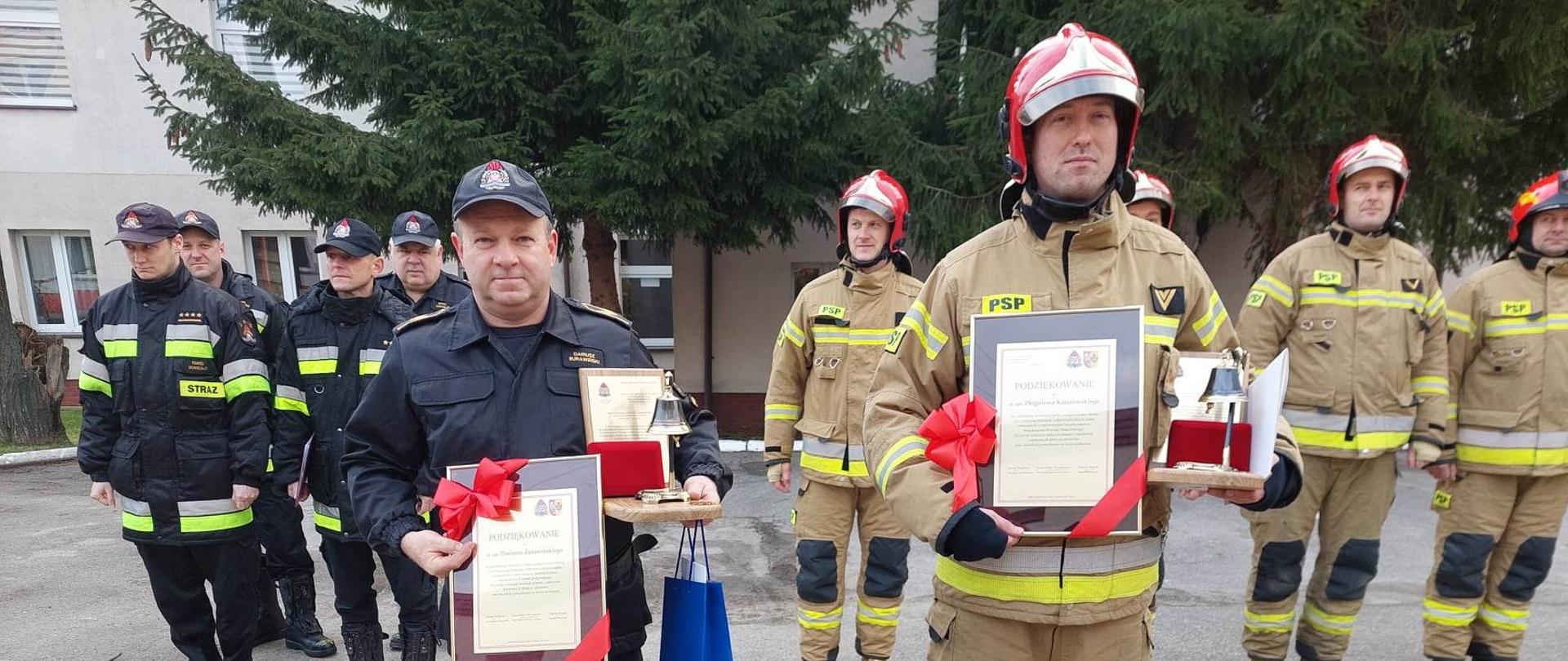 Uroczysta zmiana służby na placu Komendy. Dwie zmiany służbowe, funkcjonariusze Komendy oraz zaproszeni goście stoją naprzeciwko siebie. Po środku stoi dwóch funkcjonariuszy, w rękach trzymają upominki.