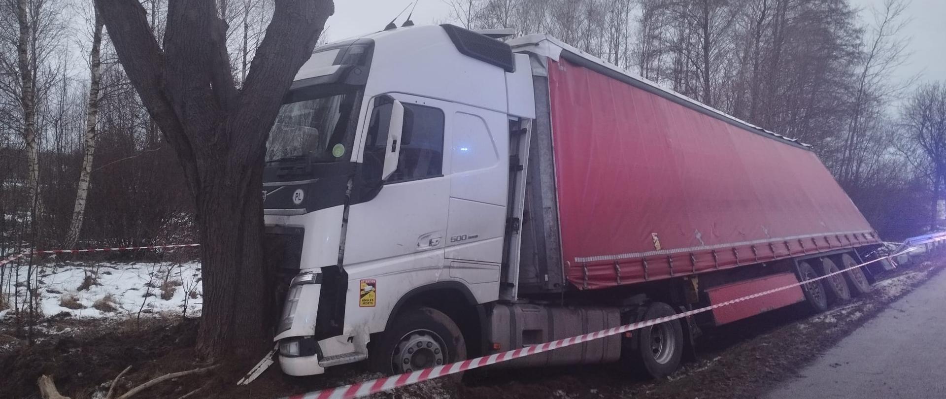 Zdjęcie przedstawia ciągnik siodłowy Volvo wraz z naczepą który uderzył czołowo w przydrożne drzewo. Kabina oparta o drzewo, a naczepa mocno pochylona stoi w przydrożnym rowie. Auto wydzielone taśmą ostrzegawczą