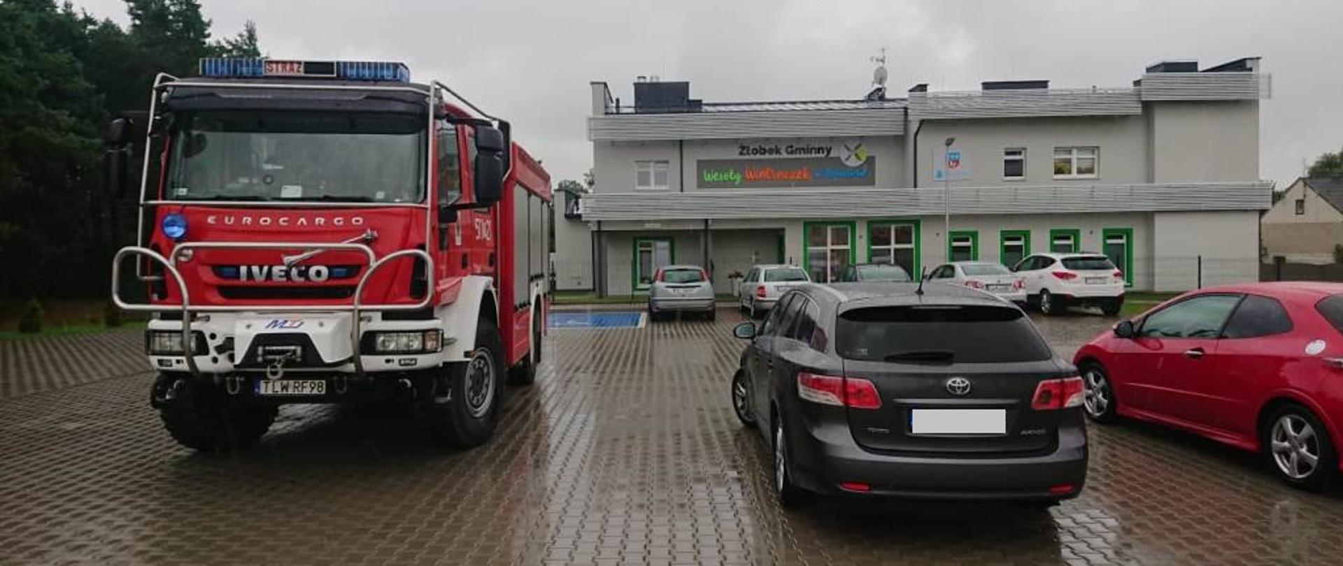 Parking przed obiektem żłobka, samochód pożarniczy Iveco oraz obok samochody osobowe, na drugim palnie budynek Żłobka Gminnego „Wesoły Wiatraczek”