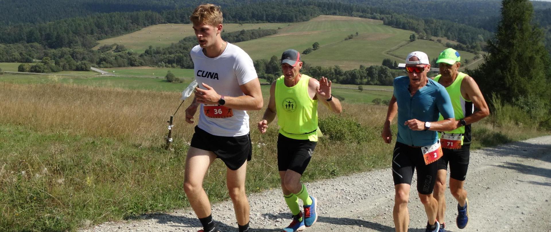 Górska – szutrowa – droga. Na niej czterech, ubranych w sportowe stroje mężczyzn, w trakcie biegu. 