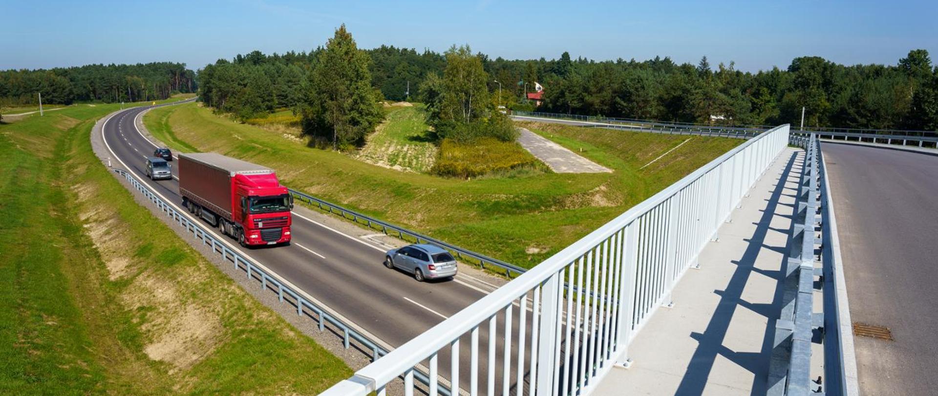 Zdjęcie wykonane z wiaduktu nad drogą krajową jednojezdniową. Na drodze ruch pojazdów osobowych i ciężarowych. W tle tereny leśne.