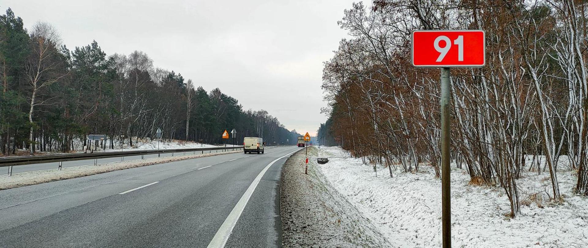 Fotografia z poziomu terenu dwujezdniowej drogi krajowej nr 91 w aurze zimowej. Droga przebiega z łuku poziomym przez obszary leśne. Na jezdniach drugi poruszaj się pojazdy. W pasie rozdziału oraz na poboczach widoczna cienka warstwa śniegu