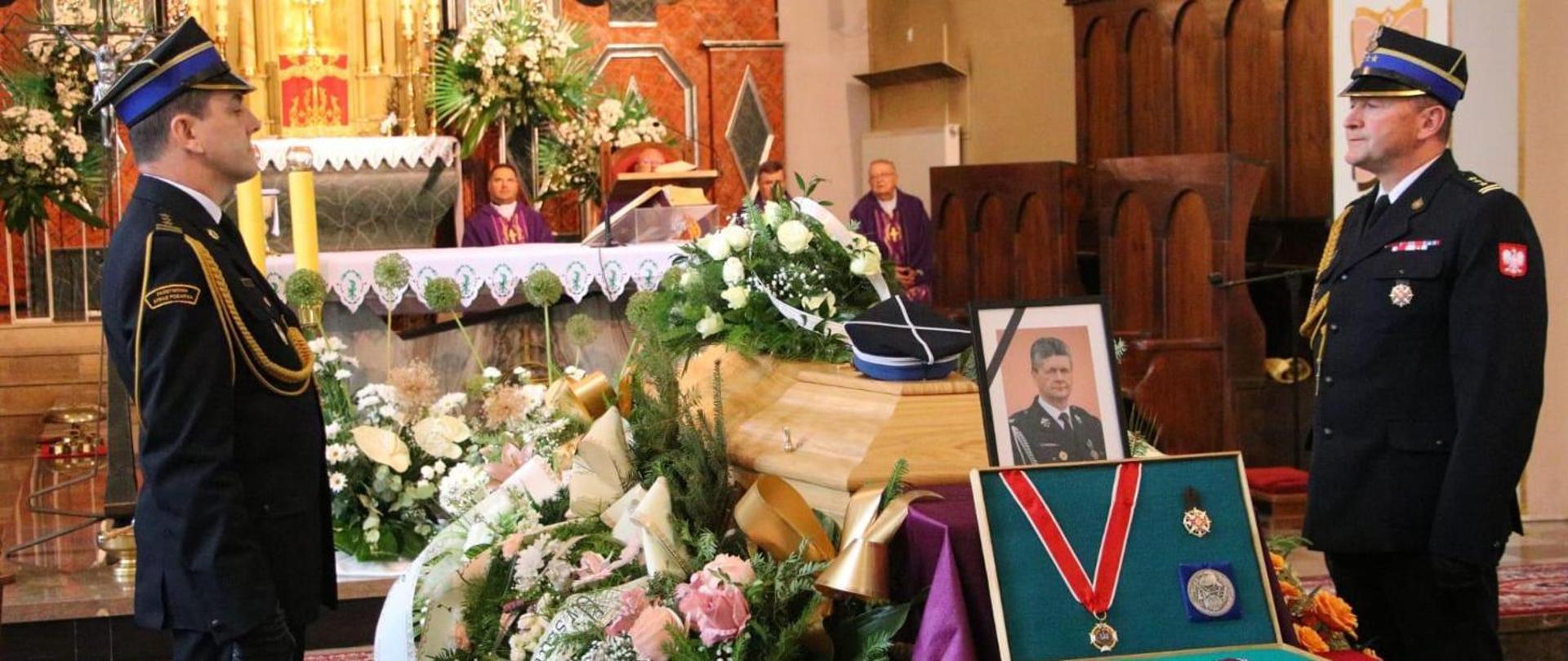 Kolorowa fotografia wykonana wewnątrz kościoła. Przedstawia asystę honorową przy trumnie przez oficerów podczas pogrzebu zasłużonego strażaka PSP i OSP. Przy trumnie znajduje się zdjęcie zmarłego strażaka i odznaczenia.