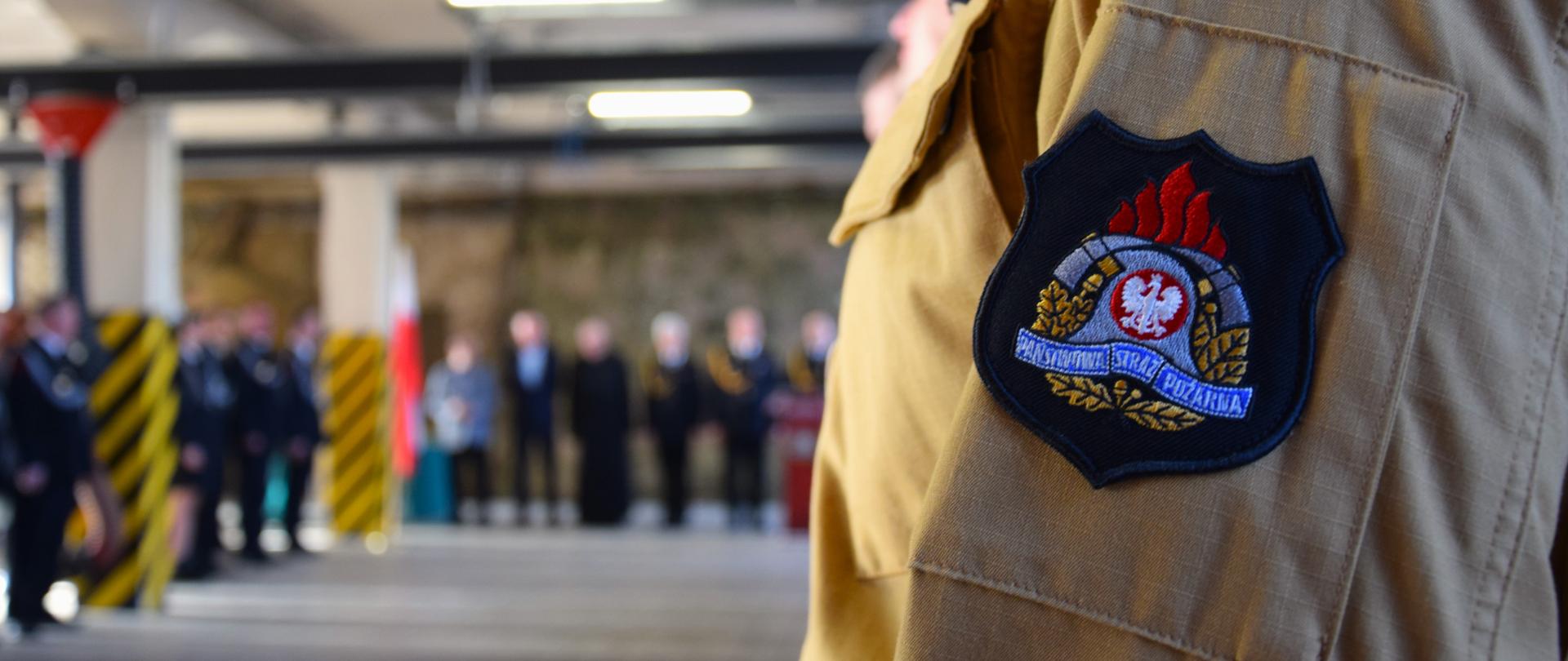 Na pierwszym planie logo Państwowej Straży Pożarnej i polska flaga naszyte na rękawie munduru funkcjonariusza. W tle uroczysta zbiórka i zaproszeni goście (rozmazane)