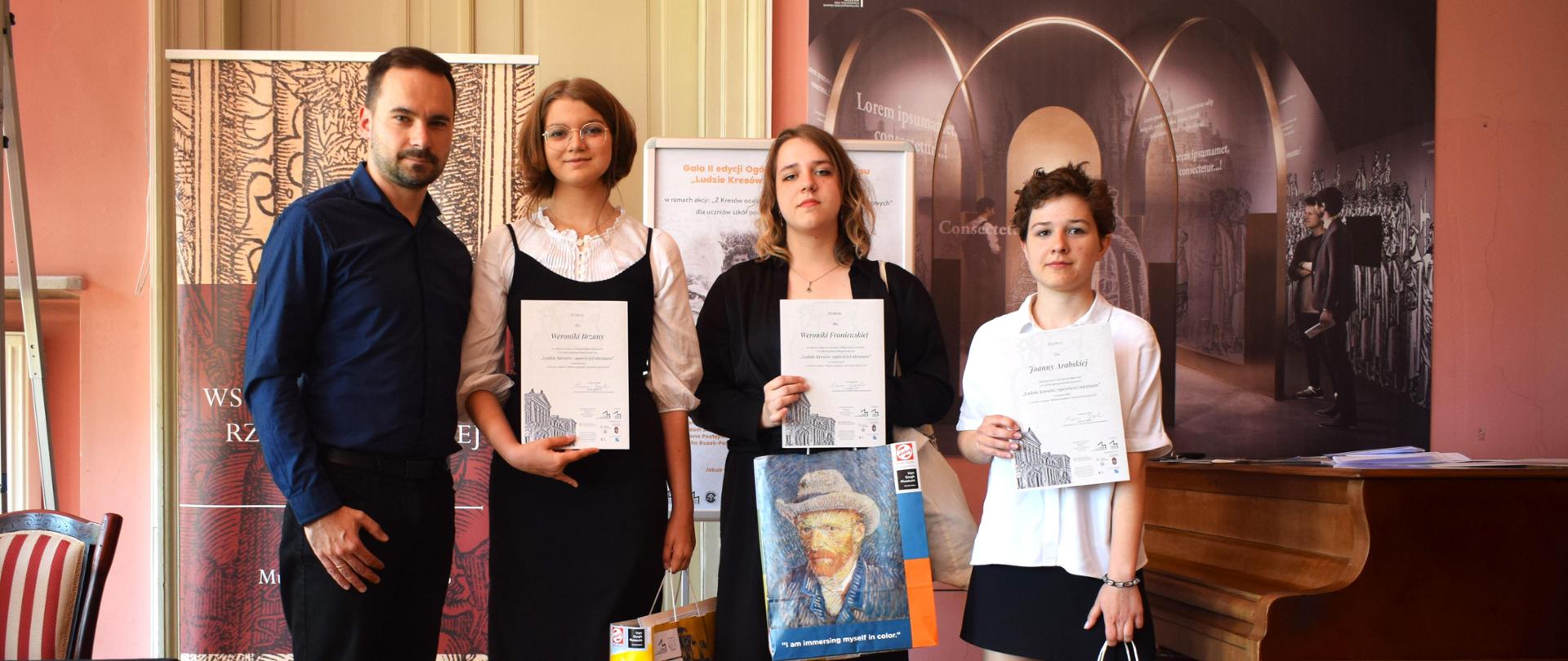 W Pałacu Potockich w Lublinie stoją nauczyciel i uczennice, które pokazują dyplomy. W dugiej ręce trzymają papierowe torby z prezentami.