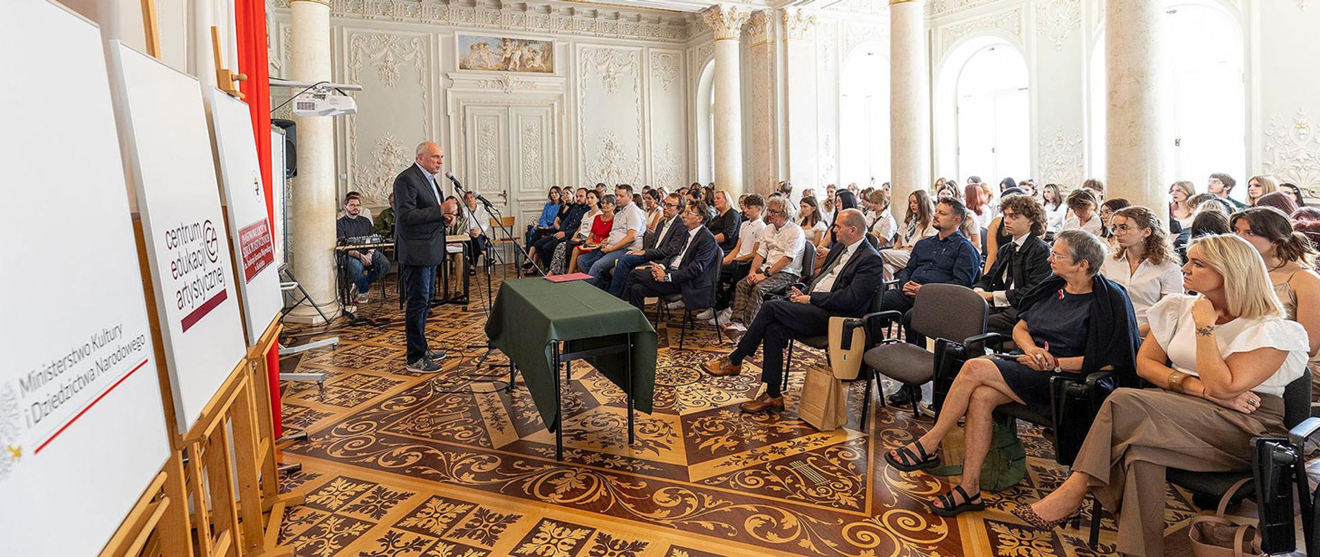 W eleganckim pomieszczeniu z wysokim sufitem i zdobną podłogą odbywa się uroczystość z okazji rozpoczęcia roku szkolnego. Po lewej stronie, w dobrze skrojonym garniturze, stoi mężczyzna i przemawia do zgromadzonych. Trzyma w ręku kartkę z przemówieniem. Obok niego stoi wysoki stolik nakryty ciemnozielonym obrusem, na którym leży księga i stoi wazon z białymi i czerwonymi kwiatami. Po prawej stronie od mówcy siedzi na krzesłach liczna grupa osób - dorośli w różnym wieku, zarówno kobiety, jak i mężczyźni. Ubrani są schludnie, niektórzy w koszule, inni w bluzkach. Wszyscy patrzą w kierunku mówcy, słuchając z uwagą. Atmosfera jest podniosła i uroczysta.