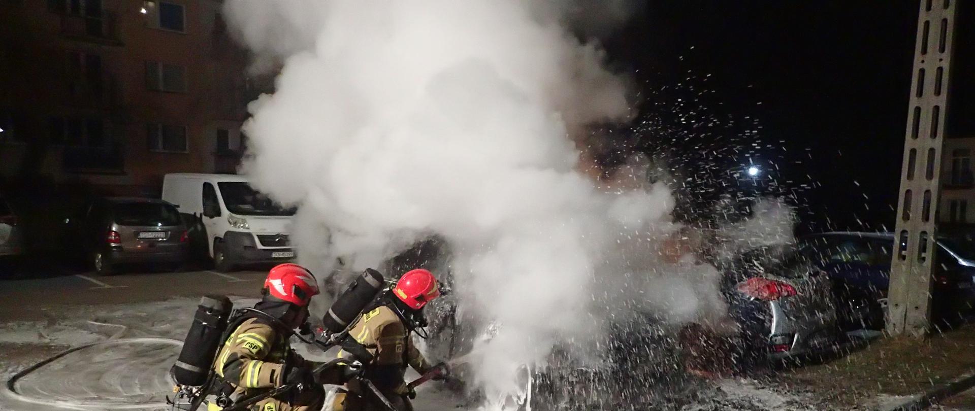 Dwóch ratowników podaje prąd piany ciężkiej na palący się samochód, dużo dymu pora nocna