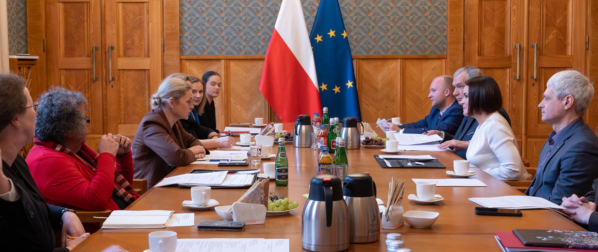 Duża sala ze ścianami w zielony wzorek, przy drewnianym stole siedzi kilkanaście elegancko ubranych osób. Po prawej stronie delegacka Białorusi po lewej kierownictwo MEN. Na stole rozłożone notatki oraz ustawione napoje. W tle flagi Polski i UE