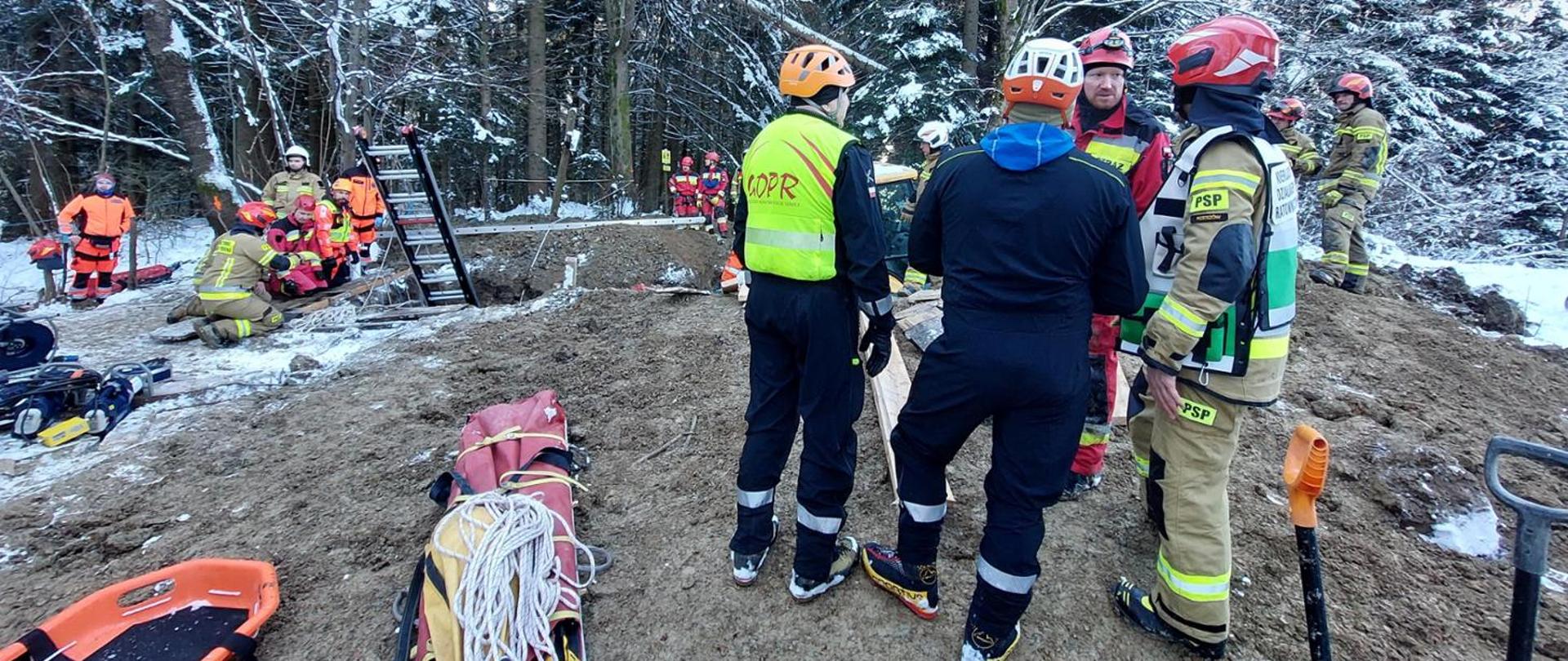 Kolorowa fotografia wykonana na zewnątrz. Przedstawia ratowników różnych służb przy wykopie w którym znajduje się przysypany mężczyzna. Z boku składowany jest sprzęt, ratownicy w ubraniach specjalnych i hełmach, wśród nich Kierujący Działaniem Ratowniczym oznakowany kamizelką.