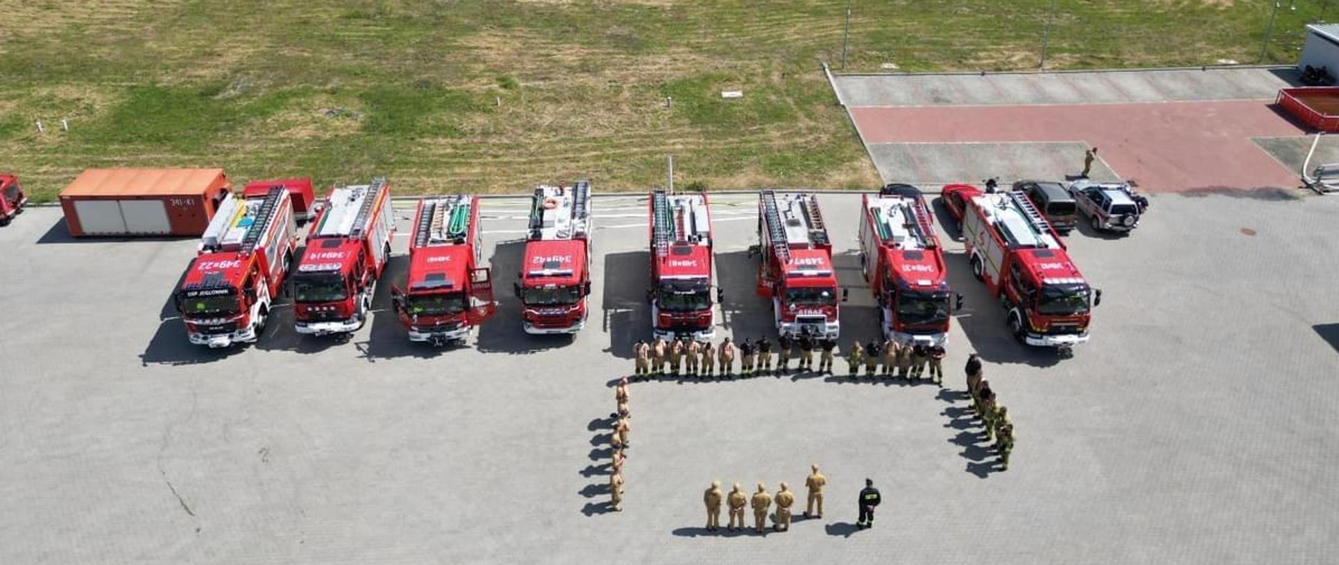 Zdjęcie zrobione z wysokości. Na placu wewnętrznym jednostki ratowniczo- gaśniczej stoją pojazdy pożarnicze. przed nimi stoją zgromadzeni funkcjonariusze. W tle widać budynki mieszkalne.