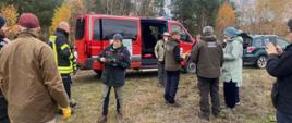 na pierwszym planie stoi grupa osób, uczestników spotkania, w wiekszości odwrócona plecami do aparatu. na drugim planie samochód strażacki typu bus. wszystko dzieje się na jakieś polanie leśnej.