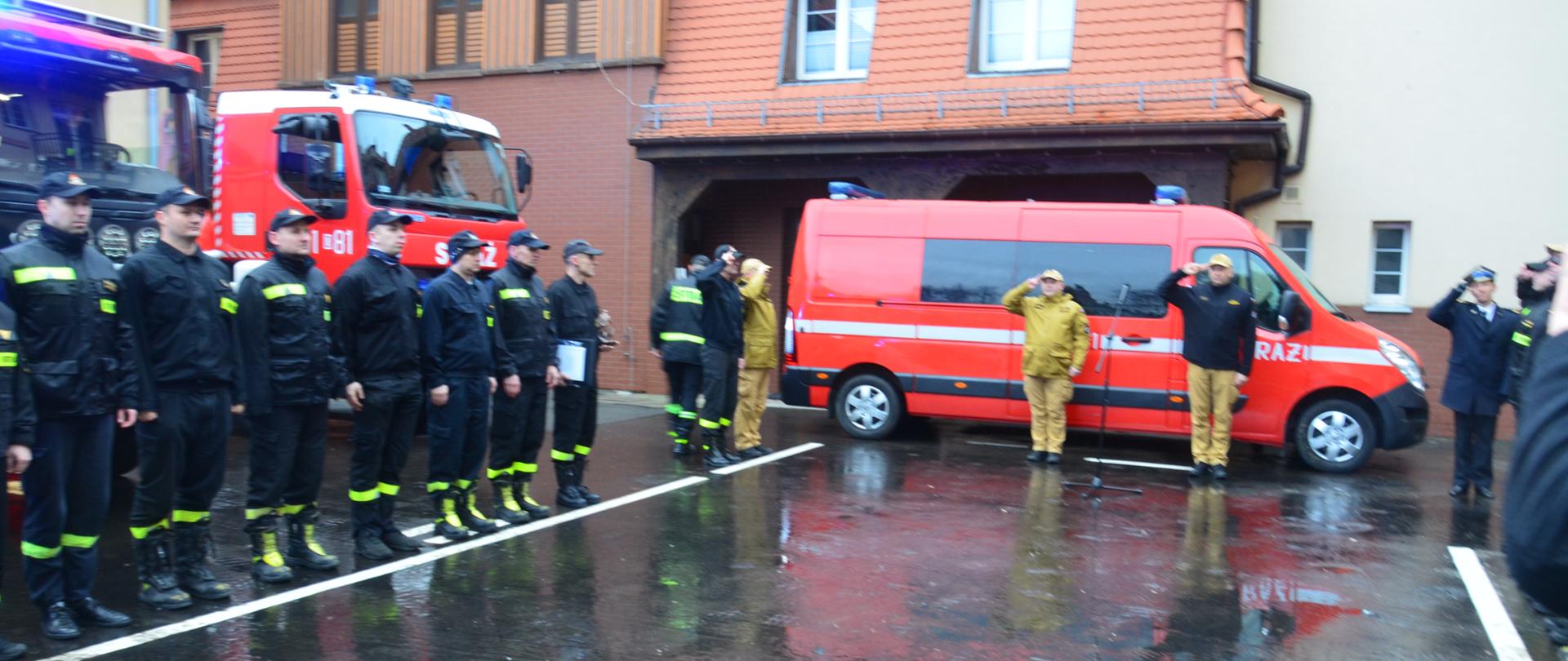 Uroczysta zmiana służby na placu Jednostki Ratowniczo-Gaśniczej nr 1. Strażacy stoją na baczność, w tle czerwone samochody strażackie