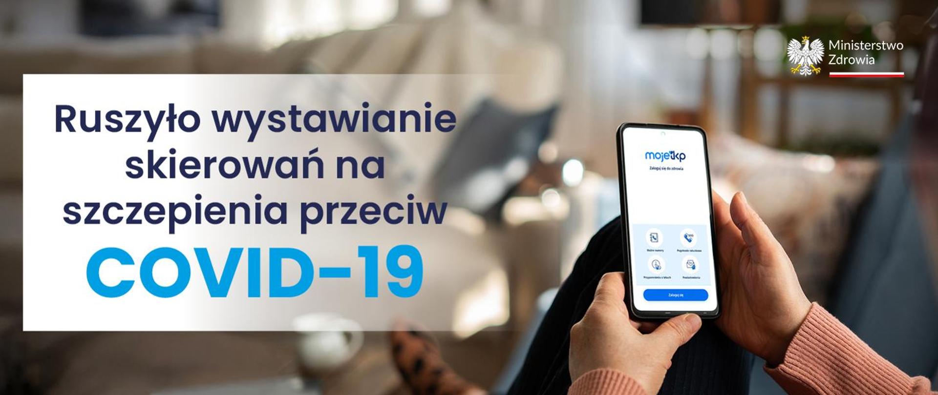 Baner reklamujący szczepienie przeciw COVID-19. W prawym górnym rogu widoczne jest logo Ministerstwa Zdrowia. W prawej części zdjęcia widoczny jest smartfon z włączoną aplikacją mojeikp, trzymany w dłoni.