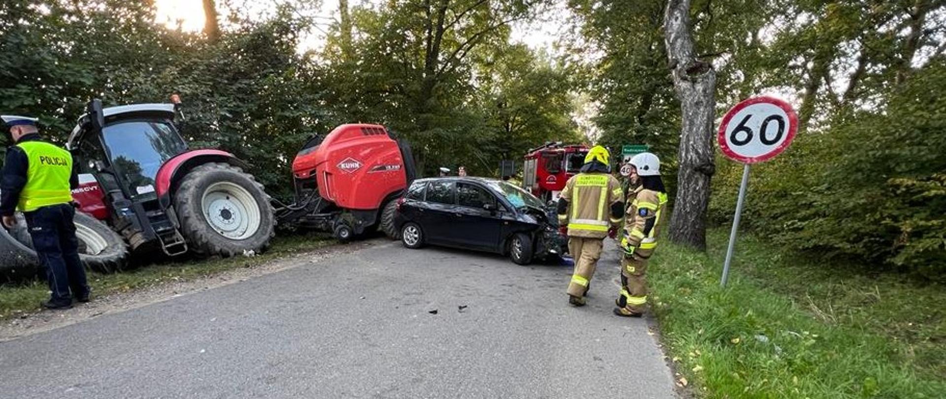 Na zdjęciu uszkodzony traktor rolniczy z czerwona przyczepą, na drodze uszkodzone czarne auto, przy nim strażacy.