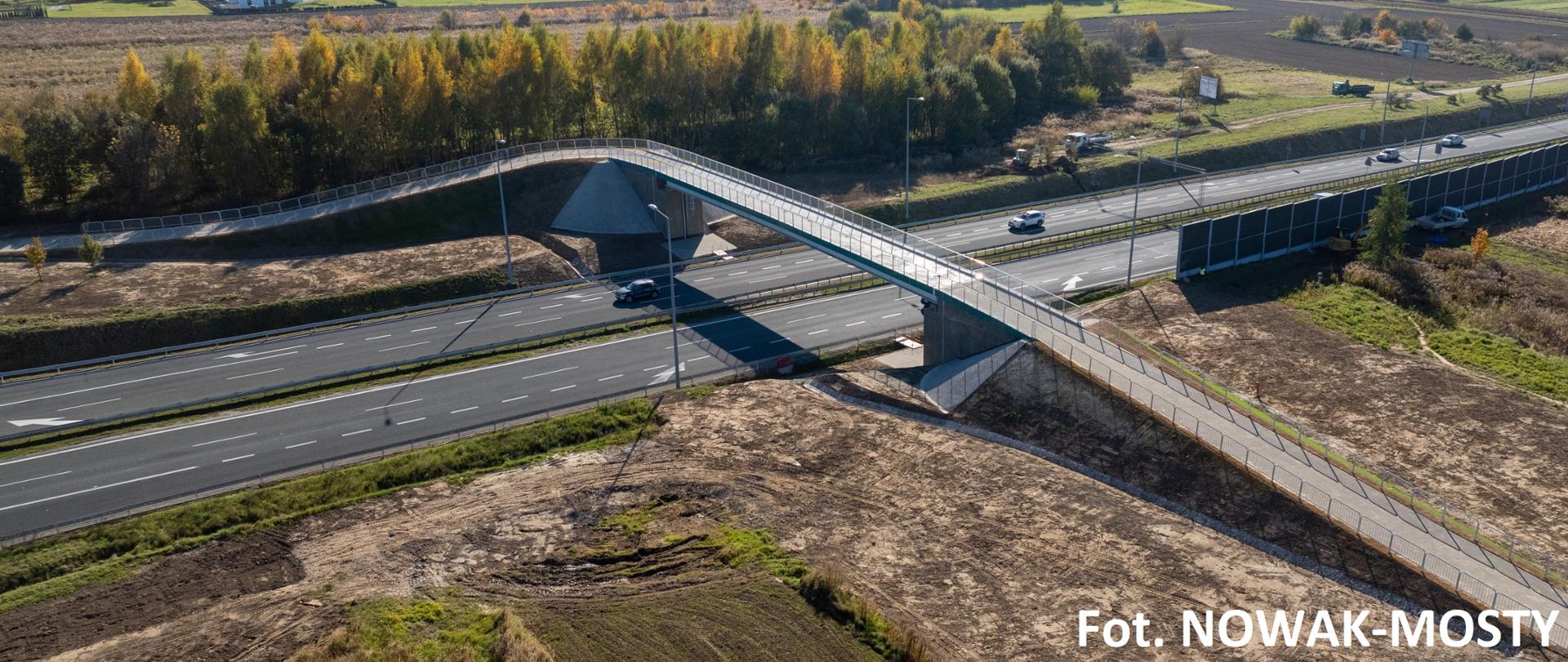 Kładka w Modlnicy, w tle pola, domy i wzgórza, kładka biegnie na DK94