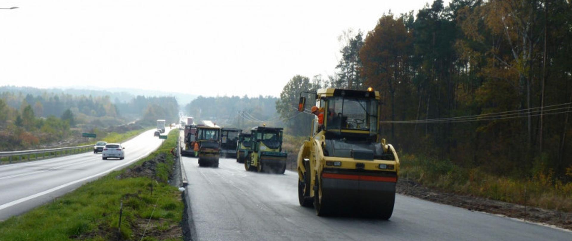 Zdjęcie z budowy drogi. Po prawej stronie znajduje się kilka ciężkich maszyn, w tym walec drogowy, a droga wydaje się być świeżo wyasfaltowana. Po lewej stronie drogi widać ruch samochodowy, który omija obszar budowy.