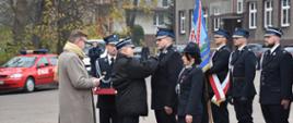 Moment wręczania odznaczeń, z prawej strony poczet sztandarowy. W tle widoczne samochody. 