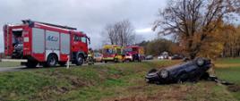 Samochód na dachu leżący na poboczu drogi, na jezdni pojazdy strażackie oraz Pogotowie Ratunkowe