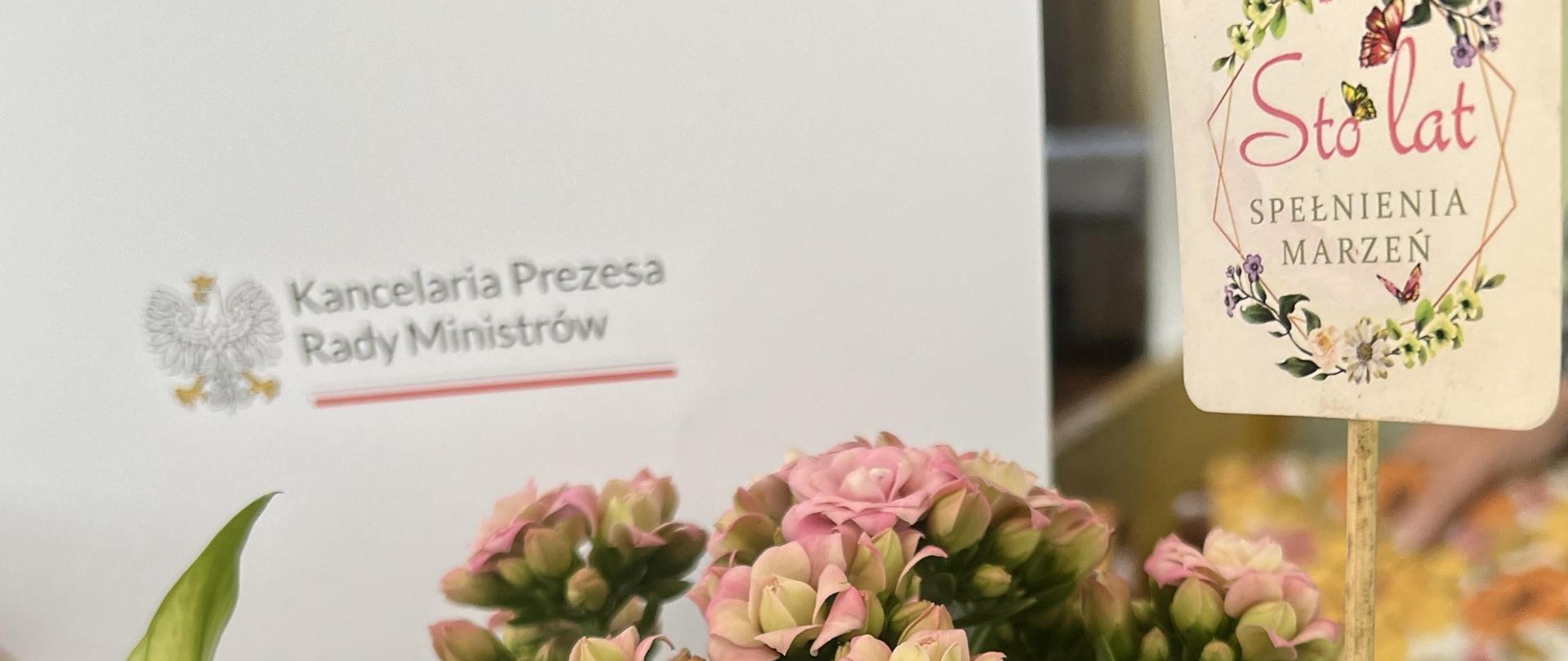 Na zdjęciu kwiaty, toper z napisem sto lat oraz teczka z listem gratulacyjnym od Premiera RP