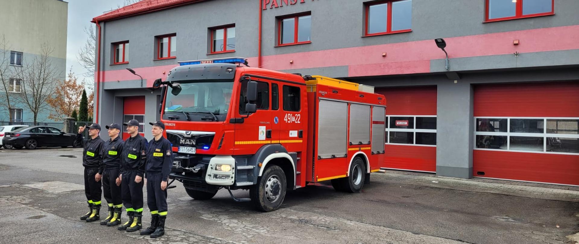 Na tle szarego budynku z czerwonymi bramami, oknami i dachem Komendy Powiatowej Państwowej Straży Pożarnej w Starachowicach oraz czerwonego strażackiego wozu bojowego czterech strażaków ubranych w czarne ubrania koszarowe oddają hołd tragicznie zmarłemu ratownikowi.