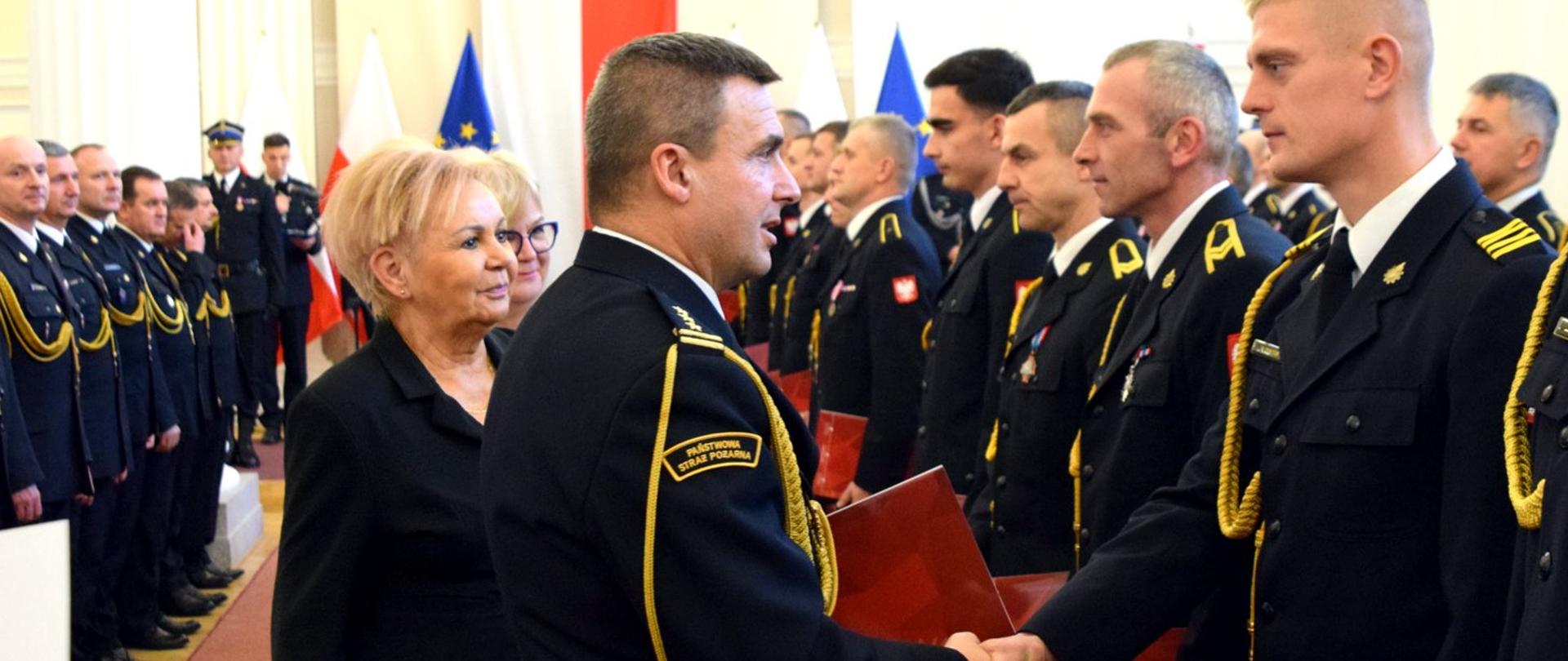 Zdjęcie zrobione w pomieszczeniu, podczas uroczystej zbiórki. Oficer Państwowej Straży Pożarnej gratuluje funkcjonariuszowi PSP awansu. W tle widać strażaków ubranych w mundury wyjściowe. Strażacy ustawieni są w szereg.