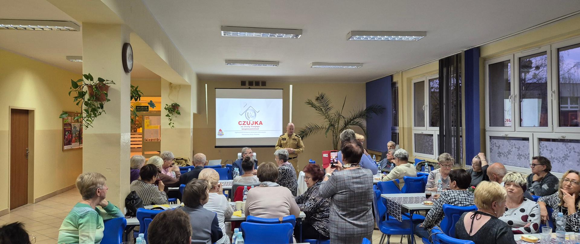 Sala w budynku. Na niebieskich krzesłach przy stolikach siedzą kobiety i mężczyźni. Skierowani są w stronę strażaka, który przedstawia prezentację.