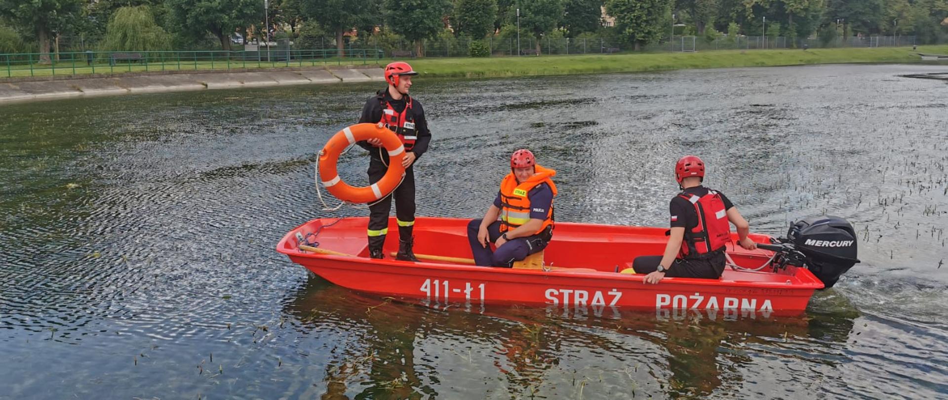 Zdjęcie przedstawia zbiornik wodny, na którym łodzią pływają strażacy wraz z policjantem zabezpieczeni w kamizelki asekuracyjne oraz kaski. Jeden z nich trzyma w rękach koło ratunkowe w kolorze pomarańczowym.