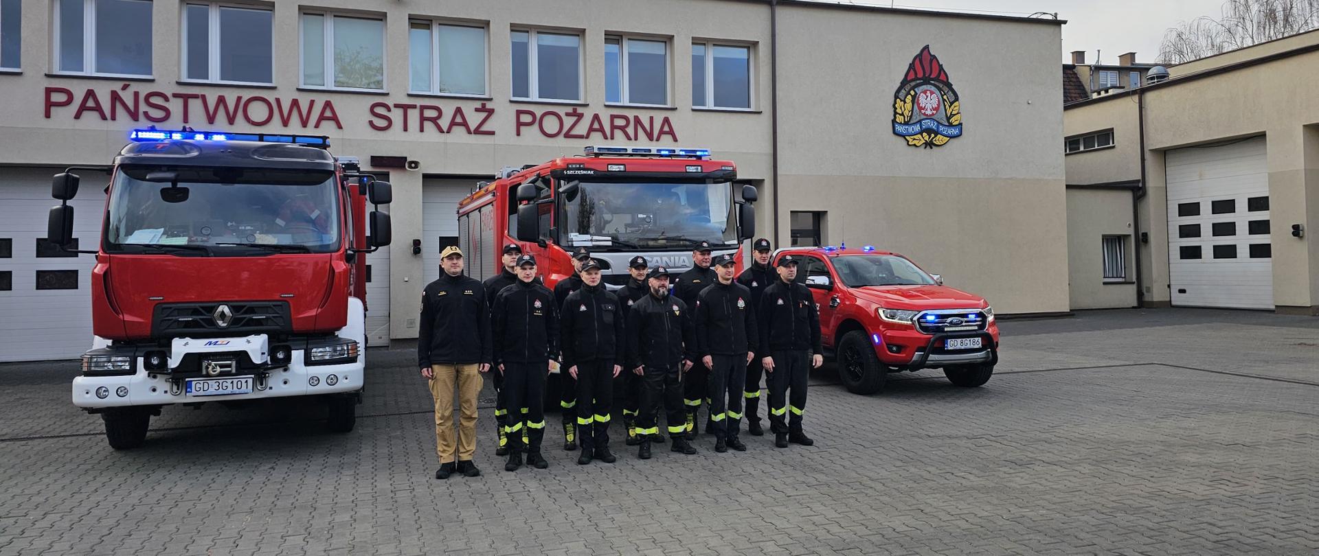 Strażacy stoją na baczność przed pojazdami i garażami oddając hołd zmarłemu.