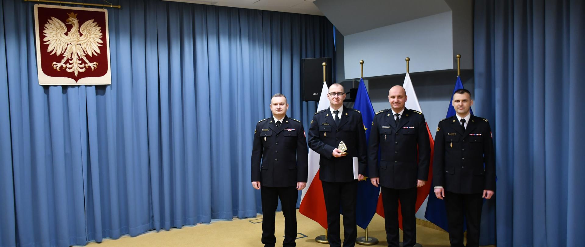 Zdjęcie przedstawia czterech strażaków w mundurach wyjściowych, stojących na tle flag Polski i UE w narożniku pomieszczenia z niebieskimi kotarami. Po lewej stronie na ścianie wisi godło Rzeczypospolitej Polskiej. 