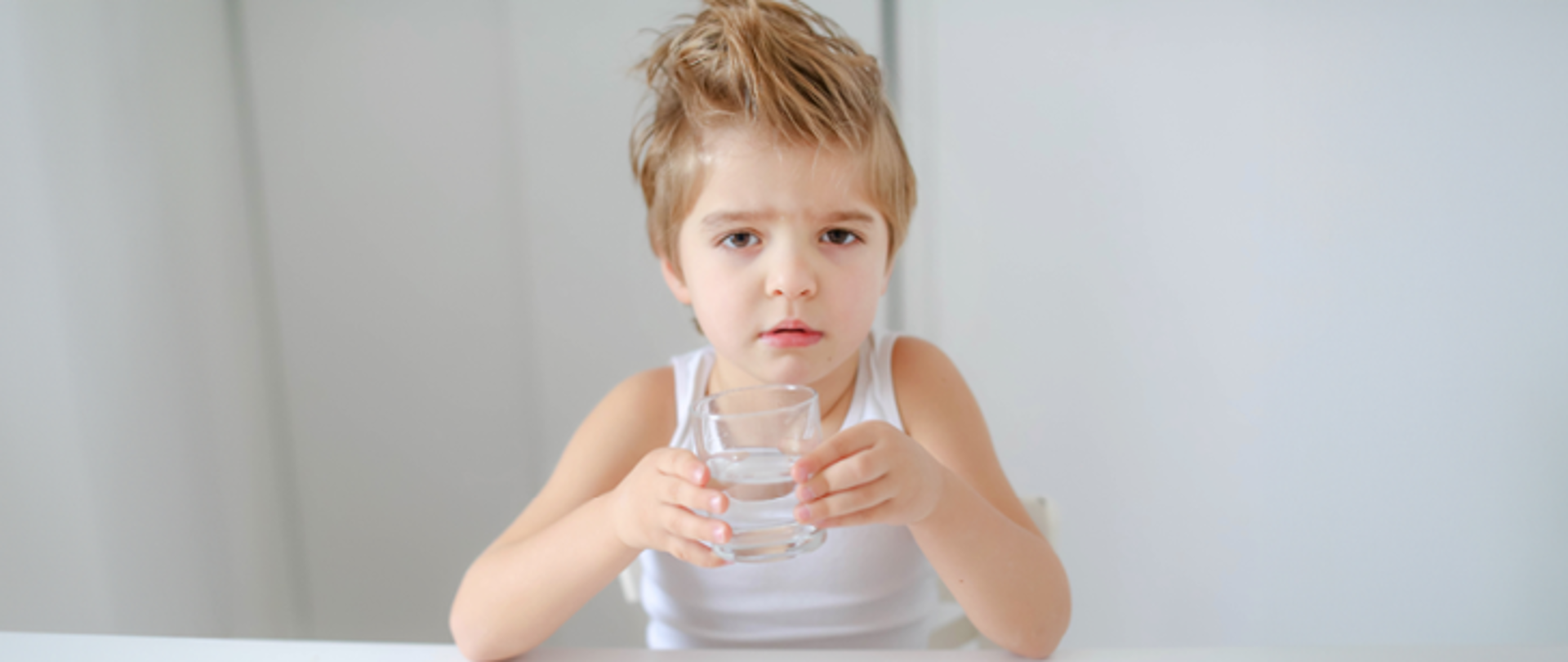Przy stoliku siedzi kilkuletni chłopiec, który trzyma w ręku szklankę z wodą 