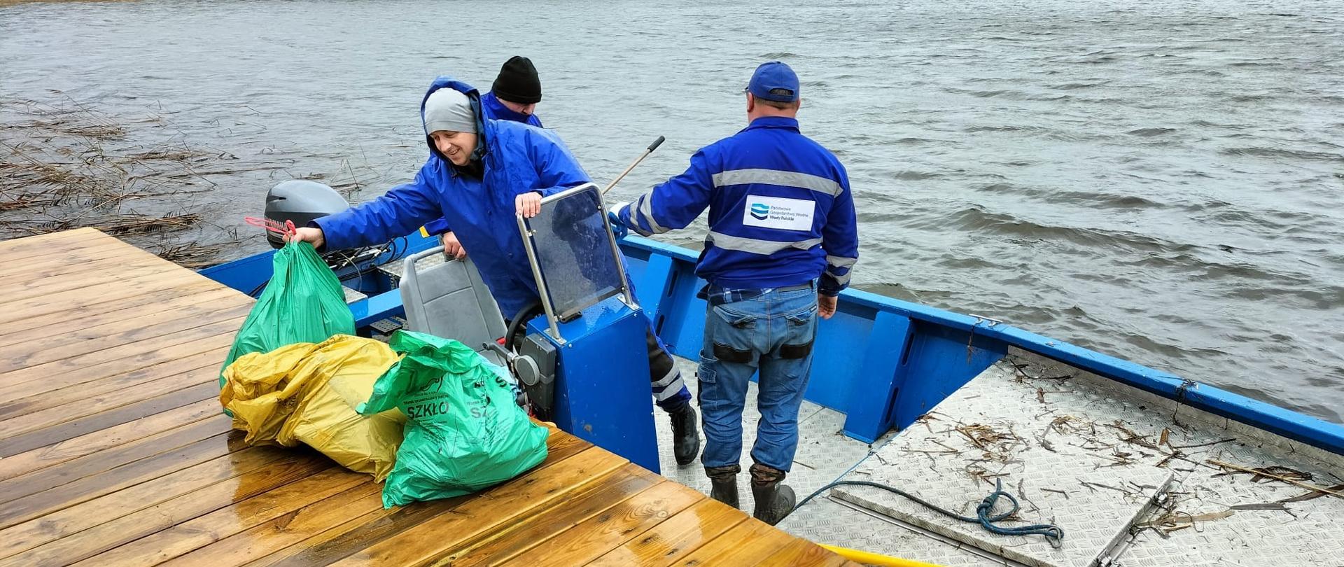 Trzech pracowników Wód Polskich stoi w łódce przy pomoście. Z łodzi wyrzucają na brzeg worki ze śmieciami. 