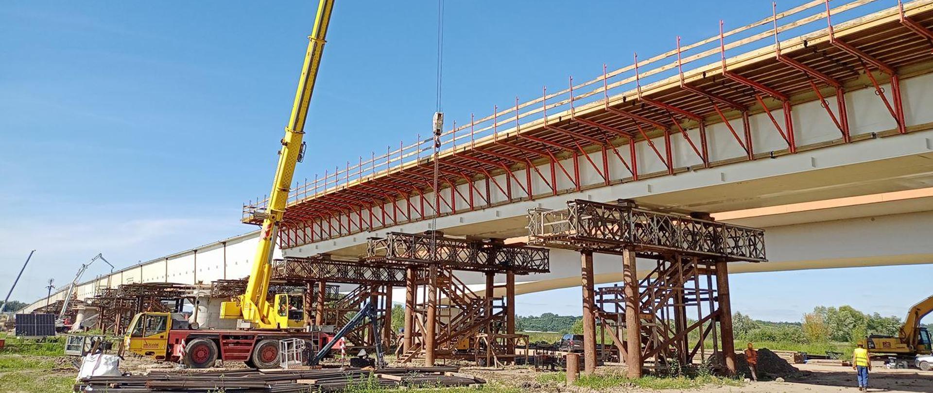 Most w Sandomierzu - konstrukcja stalowa na podporach pomocniczych. Żółty dźwig i roboty przy deskowaniu (rusztowanie wzdłuż konstrukcji) 