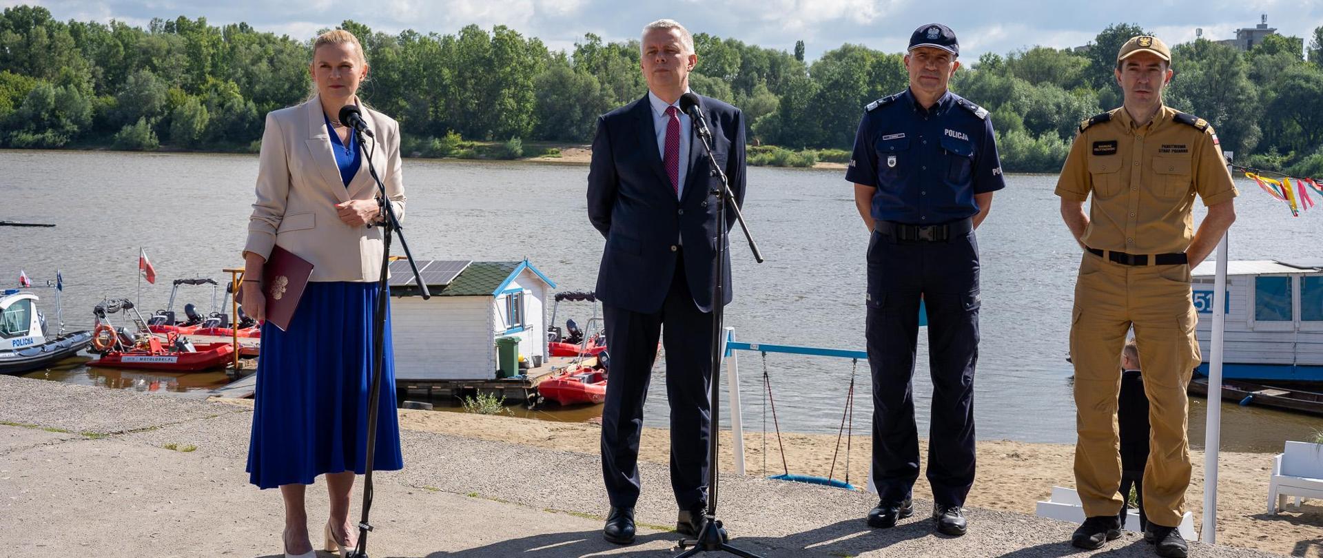 Przy dwóch mikrofonach od lewej stoją: minister edukacji Barbara Nowacka oraz szefa MSWiA Tomasz Siemoniak, następnie mężczyzna w mundurze policjanta i mężczyzna w mundurze polowym straży pożarnej. W tle widać rzekę Wisłę i łodzie patrolowe policji i wopr.