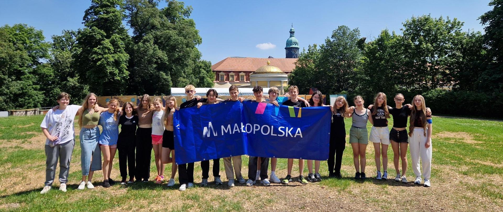 Młodzież uczestnicząca w warsztatach stoi na łące trzymając duży niebieski baner z napisem małopolska. Wszyscy stoją w rzędzie trzymając się za ramiona. W tle wysokie drzewa oraz dach i wieża zamku. 