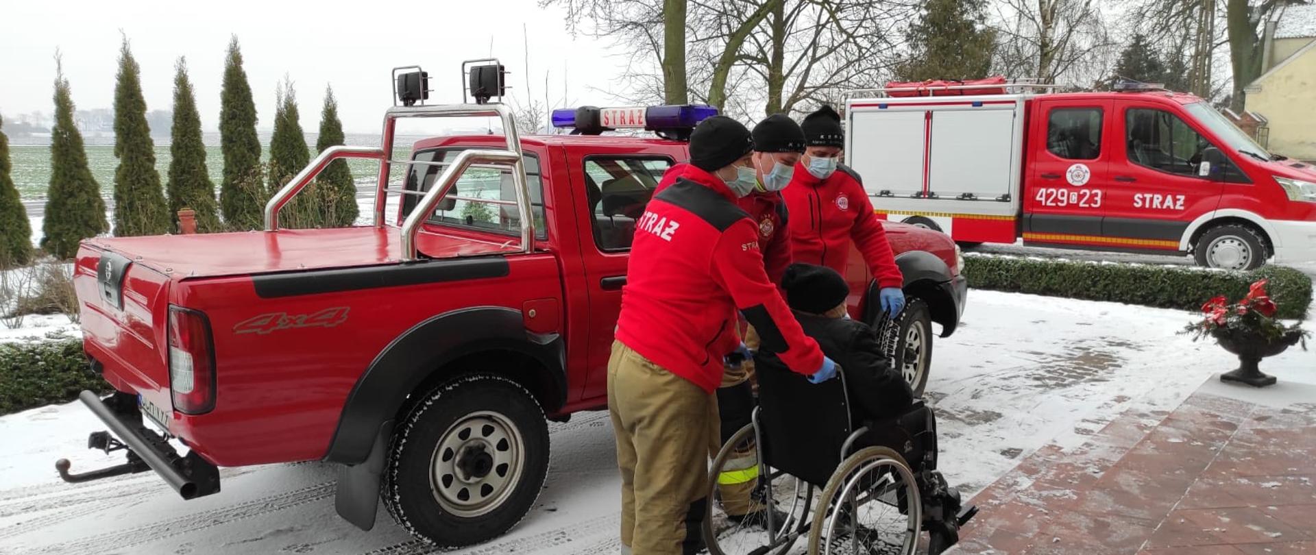 Na zdjęciu trzech strażaków przygotowuje się do wniesienia po schodach starszej osoby poruszającej się na wózku. W tle dwa pojazdy pożarnicze. Na zewnątrz panuje aura zimowa.