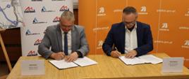 Dyrektor Oddziału GDDKiA w Opolu Rafał Pydych oraz Wiceprezes Kobylarnia S.A. Michał Niemyt podpisują umowy na budowę odcinka drogi S11. Siedzą przy stole. Oboje trzymają długopisy w prawych dłoniach i patrzą w kierunku podpisywanych dokumentów. 