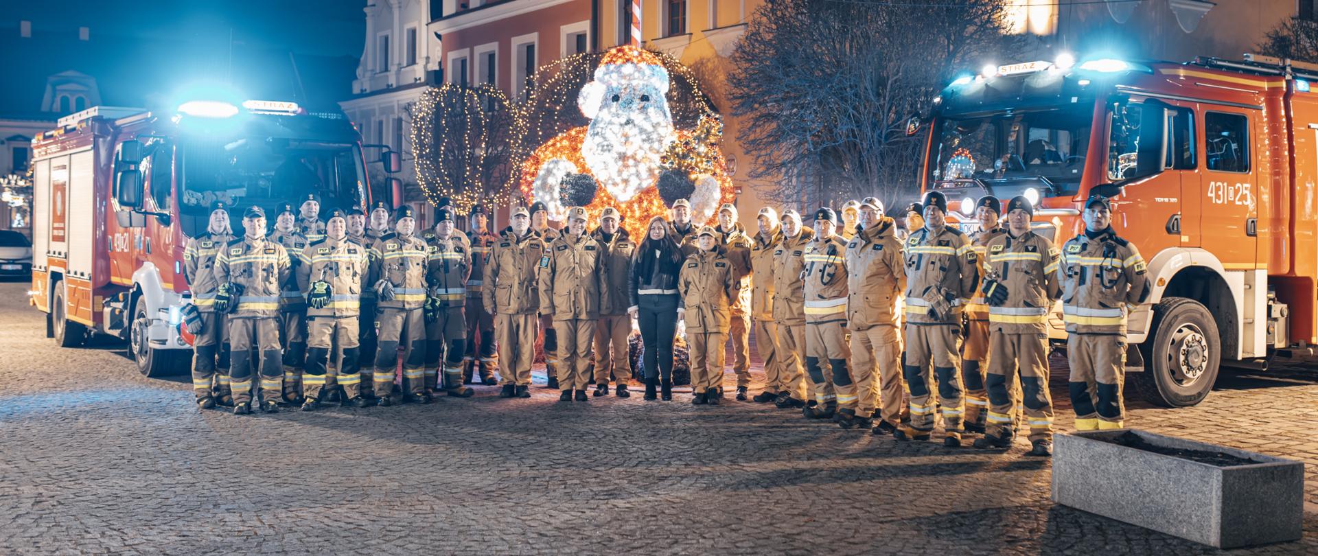 Na zdjęciu widzimy grupę strażaków w pełnym umundurowaniu stojących przed wozami strażackimi na tle pięknie oświetlonego rynku. W centralnym punkcie znajduje się dekoracja w kształcie świecącego Mikołaja, co dodaje świątecznej atmosfery. Za strażakami widoczne są zabytkowe kamienice ozdobione świątecznymi iluminacjami.