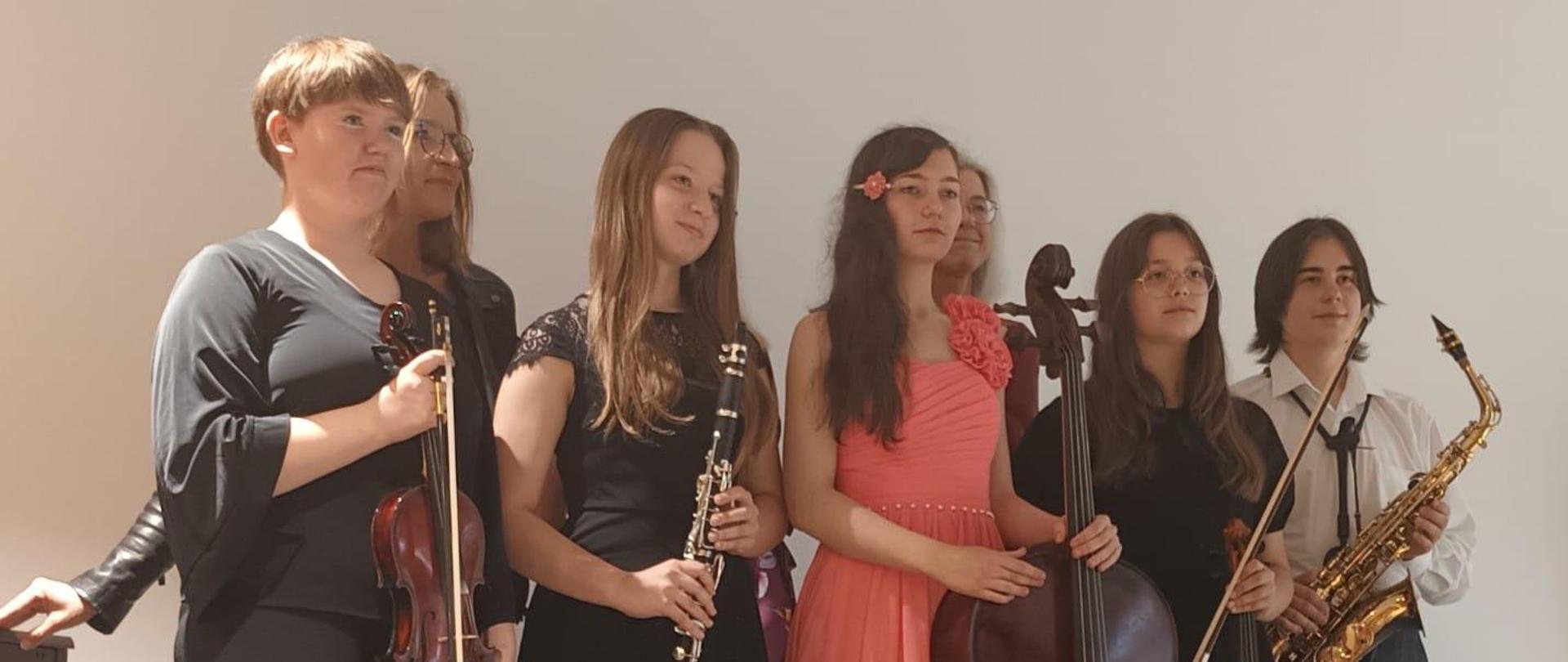 Na zdjęciu widać uczniów którzy stoją i trzymają instrumenty w rękach od lewej skrzypce klarnet wiolonczela skrzypce oraz saksofon. W tle widoczna żółta ściana