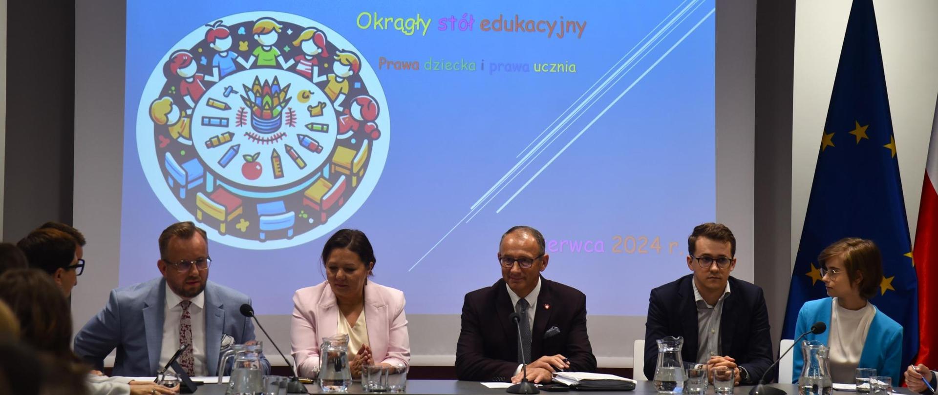 Na tle wielkiego ekranu z napisem „Okrągły stół o prawach dziecka i prawach ucznia” siedzi grupa osób, w śród nich wiceminister Izabela Ziętka.