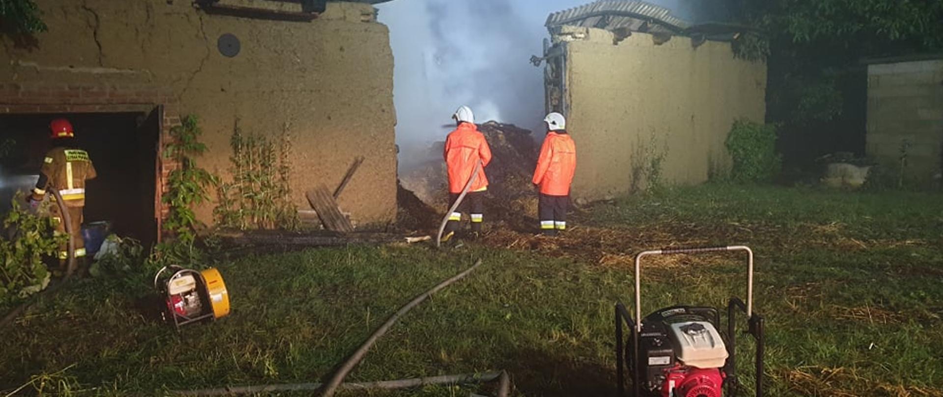 Zdjęcie przedstawia zniszczony po pożarze budynek stodoły, ściany bez dachu. Trzech strażaków dogasza wodą słomę. Przed budynkiem ustawione są dwa wentylatory oddymiające. Pora nocna.