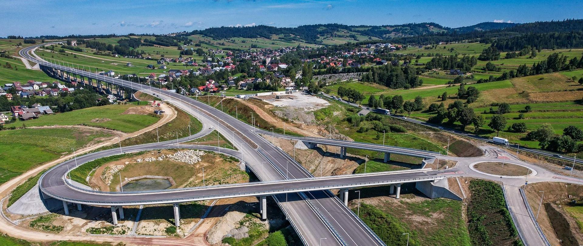 Budowany odcinek DK47 Rdzawka - Nowy Targ - na zdjęciu widoczne rondo i estakada. Po lewej stronie widać samochody poruszające się po dotychczasowym śladzie zakopianki. W oddali liczne zabudowania.