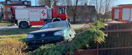 Zdjęcie przedstawia na pierwszym planie uszkodzony samochód osobowy koloru zielonego, który wbity jest w ogrodzenie posesji. Na drugim planie znajdują się dwa samochody ratowniczo-gaśnicze zaparkowane na drodze powiatowej.