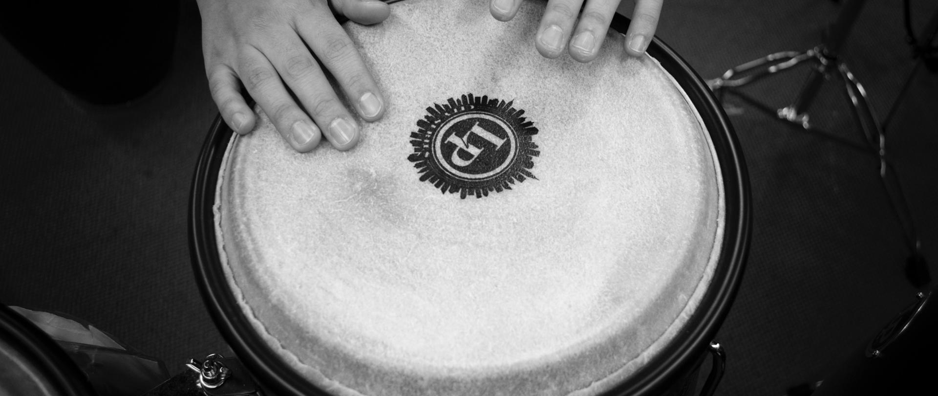 Zdjęcie przedstawia instrument perkusyjny - bęben w kolorze szarym z logo LP w kolorze czarnym , nad instrumentem znajdują się ręce osoby grającej na bębnie dookoła bębna widać zarys instrumentów perkusyjnych na ciemnoszarym tle