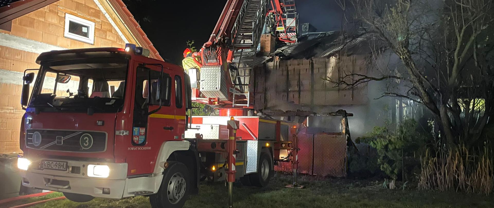 Zdjęcie przedstawia pożar budynku mieszkalnego. Jest pora nocna. Na zdjęciu widać czerwony pożarniczy pojazd typu drabina z koszem. Ramie drabiny jest rozłożone do dachu palącego się budynku. W oddali widać budynek który uległ pożarowi i wydobywa się dym z dachu. Po lewej stronie widać część budynku gospodarczego . Na ziemi znajdują się rozwinięte węże pożarnicze. Na drabinie tyłem siedzi strażak operator sprzętu ubrany w piaskowe umundurowanie specjalne na głowie ma czerwony hełm