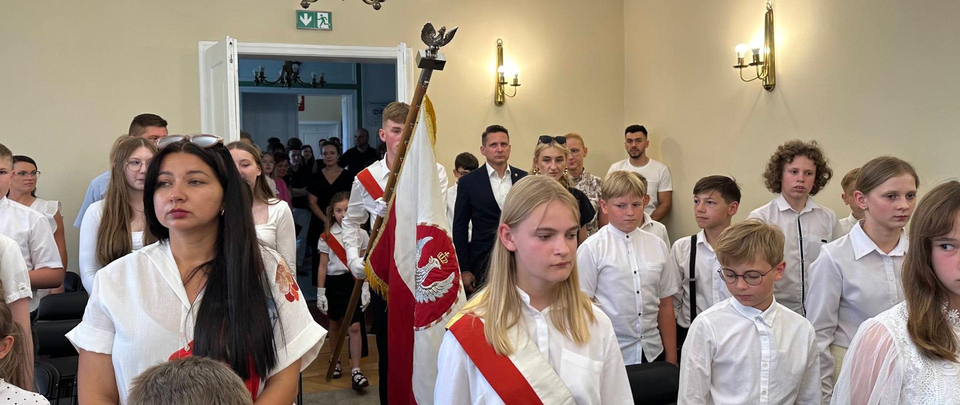 Uczniowie i rodzice stojący na baczność. Przez środek auli Poczet Sztandarowy wprowadza sztandar na uroczystość.