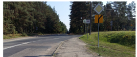 Prosta droga z rozwidleniem w prawą stronę. Po prawej stronie znak droga z pierwszeństwem pojazdu, widoczny kawałek zniszczonego chodnika. Po lewej stronie fragment lasu.